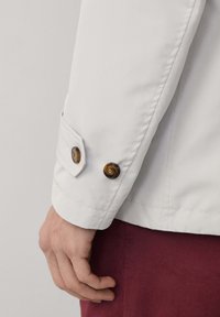 Veste blanche au design ajusté, dotée de deux boutons marron au niveau des poignets et d'une texture lisse. Le tissu est léger et frais.