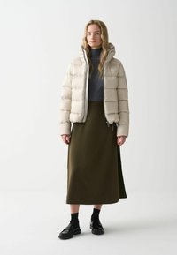 Puffjacka i ljusbeige med huva, med horisontell sömnad. Kombinerad med en lång olivgrön kjol och svarta skor.