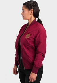 Burgundy bomber jacket made of smooth fabric, featuring ribbed cuffs, a zip closure, and a small leather patch on the left chest.