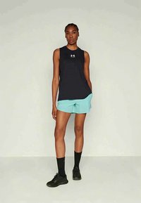 Kvinne står med hendene i lommene iført svart ärmeløs Under Armour topp, lyseblå shorts, svarte sokker og svarte joggesko.