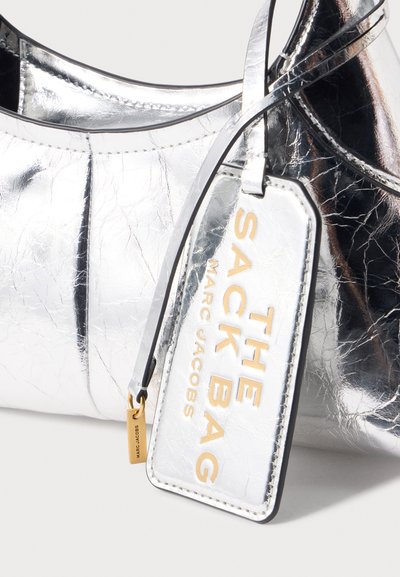 Sac à main argenté brillant Marc Jacobs avec une étiquette assortie argent et or étiquetée "THE SHACK BAG MARC JACOBS", attachée par une sangle.