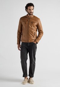 Tan læderjakke med opretstående krave, lynlås foran og to brystlommer. Båret med mørk jeans og beige slip-on sko. Glat tekstur.