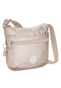 Beige crossbody bag made of smooth fabric with a zippered pocket, adjustable strap, and a charm detail. Logo patch on front.