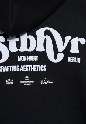 Tissu noir avec texte blanc comprenant un grand "stbhr" stylisé, des mots plus petits "Mon Habit", "Berlin", "Crafting Aesthetics", une adresse et la phrase "Worst Behavior".