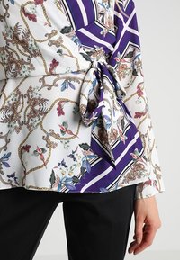 Blusa multicolore bianca con design floreali e a catena, caratterizzata da accenti viola e a righe, raccolta in vita con un fiocco.