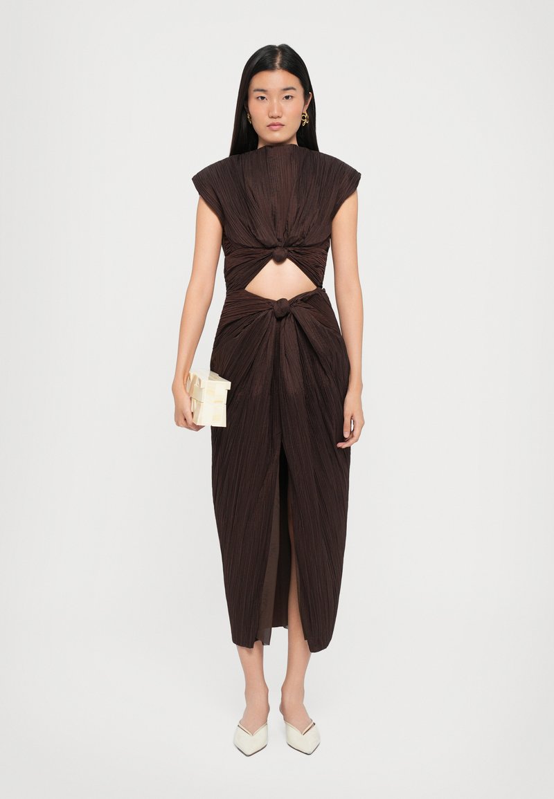 Robe plissée marron avec un col rond, des détails noués à la taille et une fente. Assortie à un petit sac à main de couleur claire.