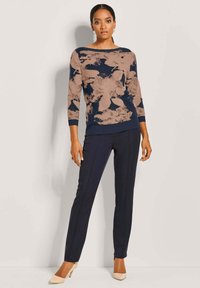 Frau in einem marineblauen und beigen, blumengemusterten Pullover, marineblauen Slim Pants und beigen High Heels, stehend vor einem schlichten hellen Hintergrund.