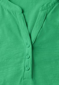 Zelený henley top vyrobený z lehkého materiálu, s výstřihem do V, čtyřmi odpovídajícími knoflíky a jednoduchým prošitím podél okrajů.