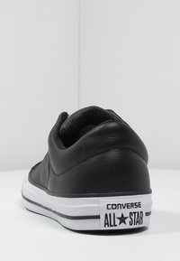 Černé kožené tenisky Converse s nízkým profilem, které mají bílé akcenty na podrážce a černobílou logo značku na patě.