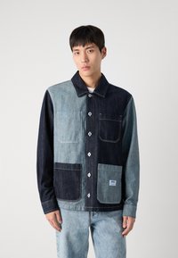 Denimjakke med patchwork-design i lyse og mørke blå nyanser, med knappelukking og flere lommer. Knappepynt på fronten.
