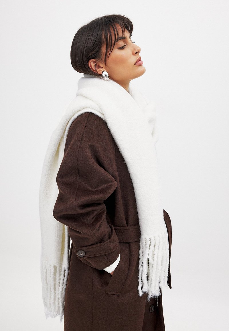 Sciarpa di lana bianca posata su un cappotto marrone, con estremità frangiate e una consistenza morbida. Il cappotto presenta bottoni visibili e maniche larghe.