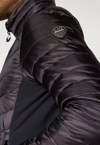 Mīkstais melnais jaka ar gludu tekstūru un horizontālām šuvēm. Tam ir izšūts logo uz piedurknes un augsts apkakles dizains.