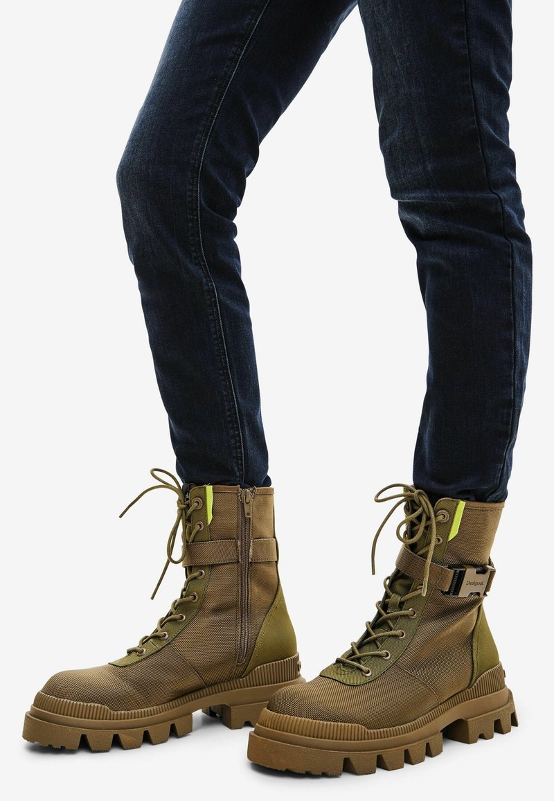 Personne portant des bottes de combat marron à lacets avec des semelles épaisses et un jean slim bleu foncé sur fond blanc.