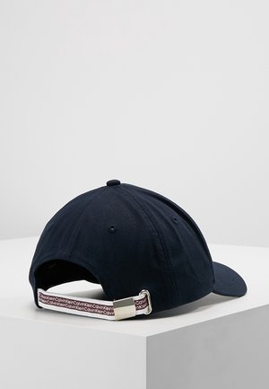 Navyblå baseballcap med justerbar stofrem med gentagende "Calvin Klein" tekst og sølvmetalspænde på hvid overflade.