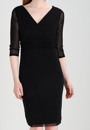 Kvinde iført en tætsiddende sort kjole med 3/4-lange, gennemsigtige ærmer og v-udskæring, stående mod en ensfarvet lys baggrund.