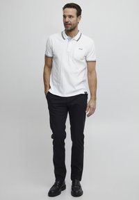Hvit polo skjorte med blå detaljer, krage med tre knapper, og brystlomme, parret med svarte bukser og svarte sko.