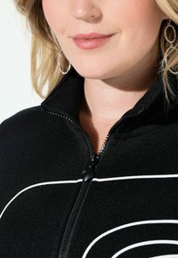 Czarna kurtka zapinana na suwak z teksturowanym wykończeniem, ozdobiona białymi, łukowatymi liniami oraz suwakiem w kształcie serca. Zbliżenie na kołnierz i zamek błyskawiczny.