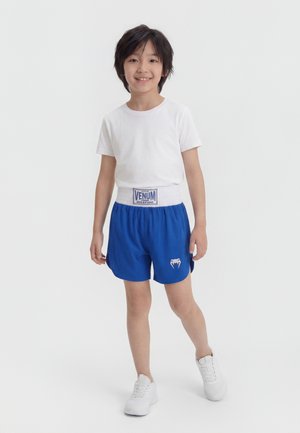 Garçon souriant debout, portant un t-shirt blanc, un short de sport bleu Venum avec logo, et des baskets blanches sur un fond clair uni.