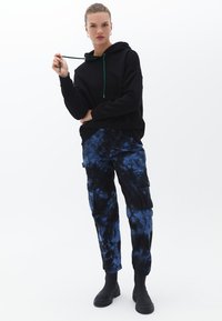 Felpa nera con cappuccio e lacci verdi, abbinata a pantaloni cargo tie-dye blu e neri. Il modello indossa stivaletti neri.