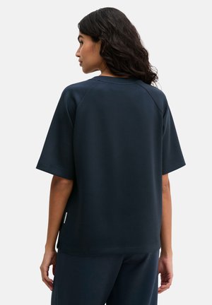 Kobieta o ciemnych, falowanych włosach, ubrana w luźną granatową bluzkę z krótkim rękawem i dopasowane spodnie, stoi tyłem na jednolitym tle.