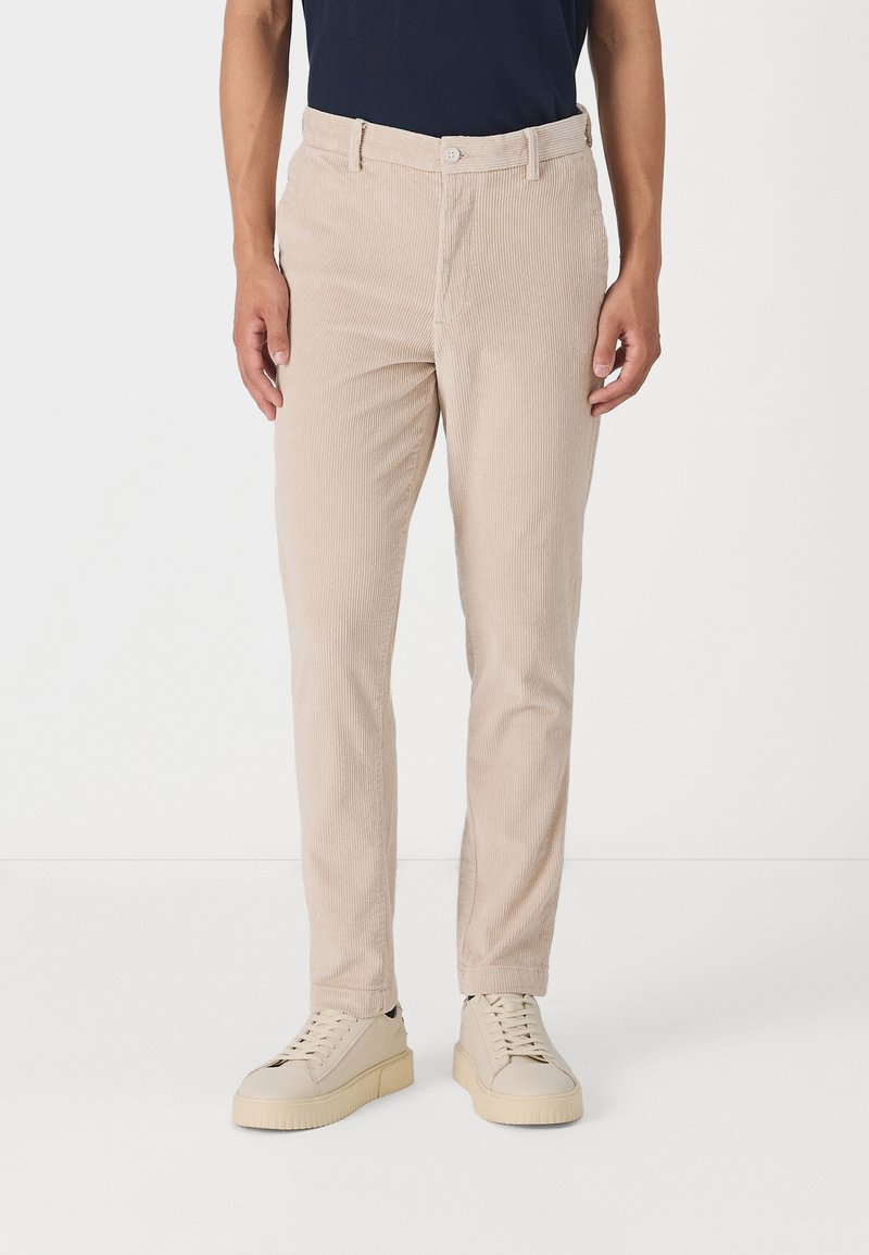 Homme portant un pantalon en velours côtelé beige slim-fit, des baskets lacées beige clair et une chemise bleu foncé, debout devant un fond blanc.