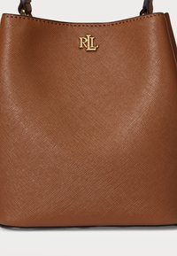 Bolsa de mão castanha com uma textura rugosa e um acabamento suave, apresentando um logotipo em tom de ouro no centro superior e uma forma estruturada.