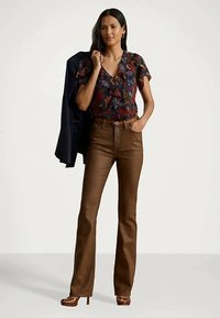 Donna che indossa una blusa a balze floreale e pantaloni in pelle marrone, mentre tiene un blazer blu navy sulla spalla, in piedi contro uno sfondo neutro.