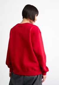 Rød sweatshirt med rund hals og senkede skuldre. Glatt tekstur, ribbede mansjetter og nederkant, vist fra baksiden med nøytral bakgrunn.