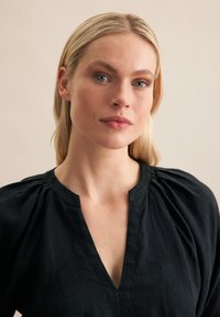 Top in lino nero con scollo a V, arricciature sulle spalle e vestibilità rilassata. La texture appare morbida e leggermente stropicciata.