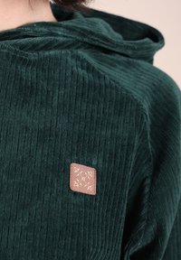 Hoodie en velours côtelé vert foncé avec des côtes horizontales, présentant une petite pièce en cuir marron avec des coutures décoratives dorées sur le côté avant gauche.