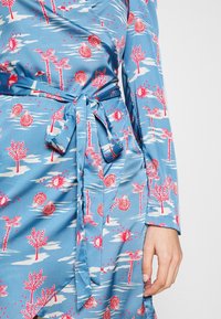 Robe portefeuille bleue avec imprimé tropical rouge et blanc présentant des palmiers, des coquillages, des étoiles et des motifs soleil, nouée à la taille avec une ceinture en tissu.
