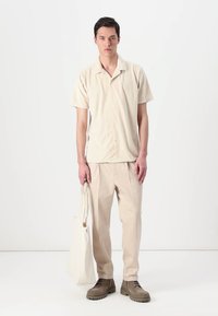 Μπεζ πουκάμισο με κουμπιά και ταιριαστά παντελόνια, συνδυασμένα με καφέ μπότες. Μια καμβάς τσάντα κρέμεται από το αριστερό χέρι. Το φόντο είναι λευκό.