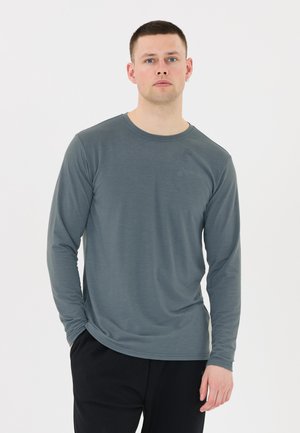 Ung mand stående med hænderne i lommerne iført en grå langærmet skjorte og sorte bukser mod en ensfarvet hvid baggrund.