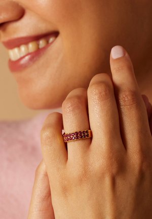 Goldring mit einem Band, das abwechselnd mit rosa und dunklen Edelsteinen besetzt ist, zeigt eine glatte Oberfläche und detaillierte Textur. Eine Hand hält den Ring.