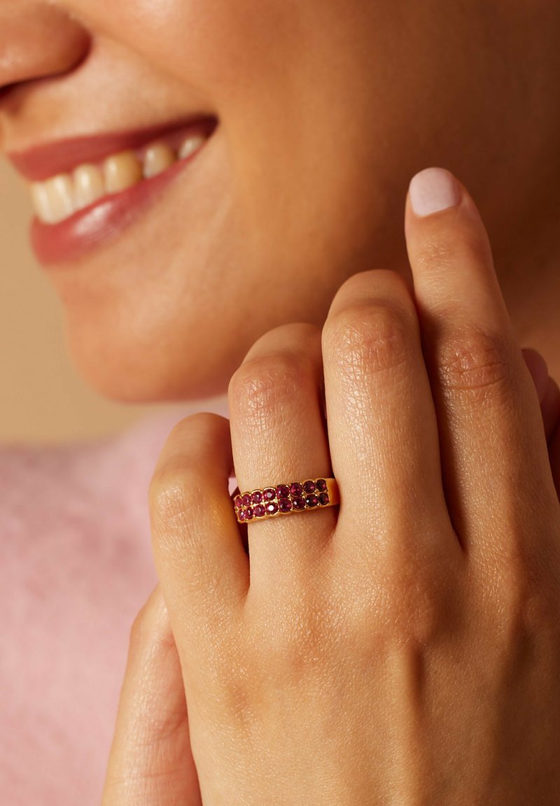 Gouden ring met een band bezet met afwisselend roze en donkere edelstenen, die een gladde oppervlakte en gedetailleerde textuur toont. Hand die de ring vasthoudt.