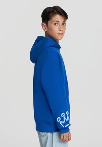 Niebieska bluza z kapturem, z kieszenią kangurką z przodu, kapturem na sznurki oraz białą grafiką korony na lewym rękawie. Wykonana z miękkiego materiału, o luźnym kroju.
