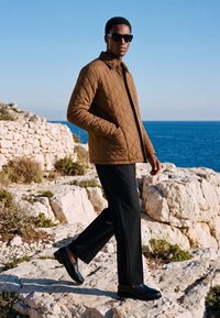 Uomo con giacca trapuntata marrone, pantaloni neri e occhiali da sole che cammina su una costa rocciosa con mare blu e cielo limpido sullo sfondo.