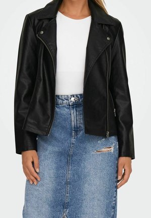 Femme portant une veste en cuir noire par-dessus un haut blanc avec une jupe en jean bleu taille haute qui a une déchirure sur le côté droit.