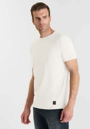 Hombre con camiseta de cuello redondo de mangas cortas blanca y jeans oscuros, de pie contra un fondo claro y liso, mirando hacia un lado.