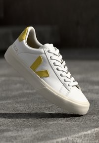 Vit lädersneaker med gula detaljer, texturerad gummisula och enkel snörningsdesign med en framträdande V-logotyp på sidan.
