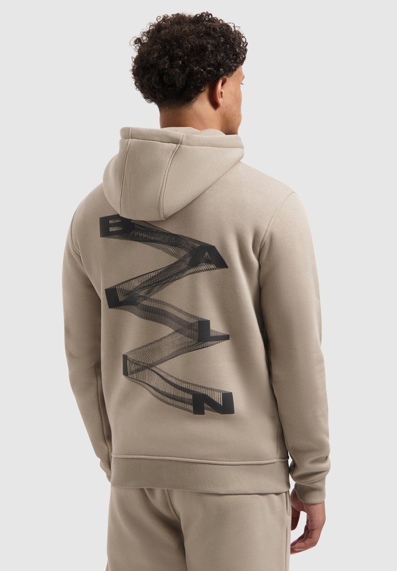 Man met krullend haar in een beige hoodie en een beige broek, met zijn rug naar de camera gekeerd. Op de hoodie staat een zwart abstract zigzag tekstontwerp met de tekst "BAALN".