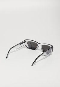 Lunettes de soleil noires et transparentes avec une forme géométrique en œil de chat, verres teintés sombres et branches fines affichant un détail de logo sur un côté.