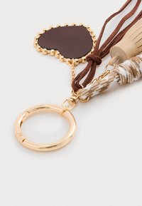 Chaveiro com um anel circular dourado, um pendente em forma de coração de couro castanho e tiras trançadas em castanho e branco com acentos decorativos.