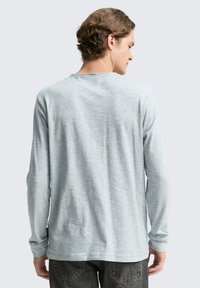 Koszula z długim rękawem w jasnoszarym kolorze, wykonana z miękkiego materiału. Posiada okrągły dekolt i luźny krój, z widoczną metką na dolnej stronie.