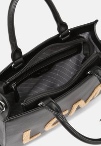 Mustakasta nahkalaukkua, jossa on teksturoitu pinta. Sisällä harmaa vuori, jossa on raidallinen kuvio, yksi vetoketjullinen tasku ja pörröinen logoyksityiskohta.