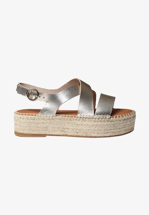 Metalické stříbrné espadrilové sandály s křížovými popruhy, nastavitelným kotníkovým sponou a tkanou juty platformou. Hladká kožená textura.