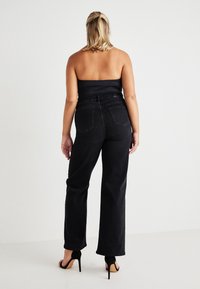 Mulher com cabelo loiro até aos ombros, vestindo um top preto sem alças, jeans pretos de cintura alta e sandálias de salto alto pretas, de costas para a câmara.