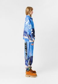 Chaqueta y pantalones de estampado azul con un gráfico de montaña, con texto blanco en la chaqueta y una etiqueta negra en los pantalones. Botas marrones.