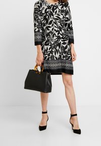 Robe noire et blanche avec des motifs de feuilles, des manches trois-quarts et une bordure noire. Tient un sac à main noir avec des poignées écaille de tortue.
