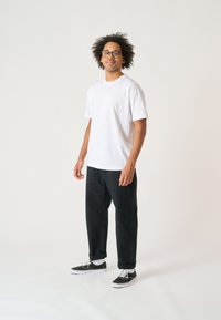 White cotton t-shirt, black straight-leg pants, and black sneakers with white soles. Model has curly hair and wears glasses.